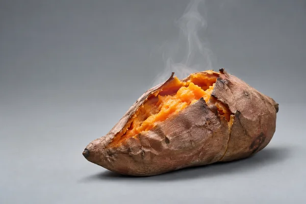 A freshly baked sweet potato with wrinkled brown skin split open to reveal bright orange flesh sits on a cool grey seamless surface, with wisps of steam rising…
