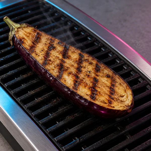 A grilled eggplant half lies diagonally cut-side up across black grill bars, with dark sear marks, a green stem and faint wisps of vapor.