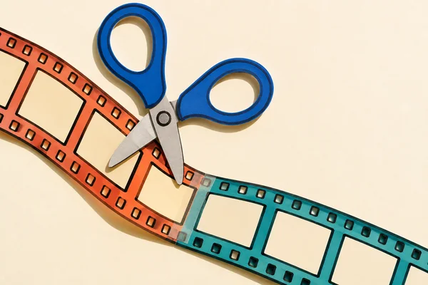 A pair of broad cobalt-handled scissors lies partly open across a curved strip of film on a cream surface.