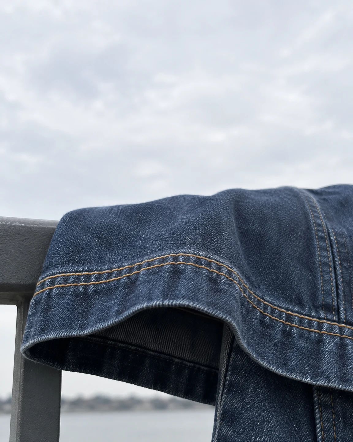 Close view of indigo denim draped over a gray outdoor railing, with two mustard-colored stitch lines curving across a folded hem beneath a pale cloudy sky.