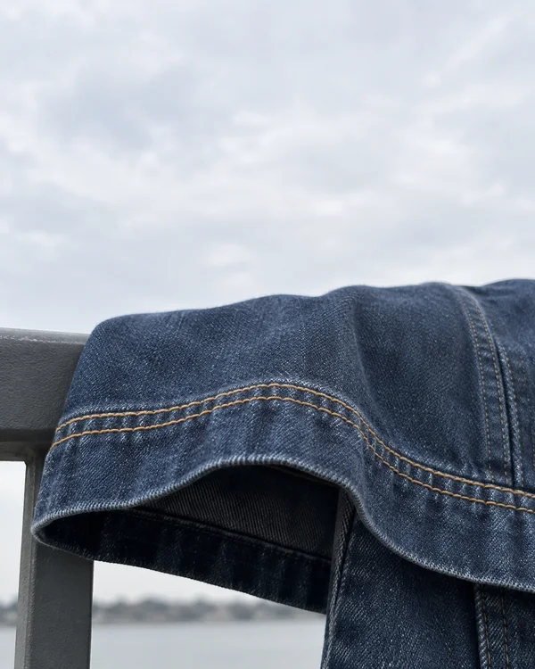 Close view of indigo denim draped over a gray outdoor railing, with two mustard-colored stitch lines curving across a folded hem beneath a pale cloudy sky.