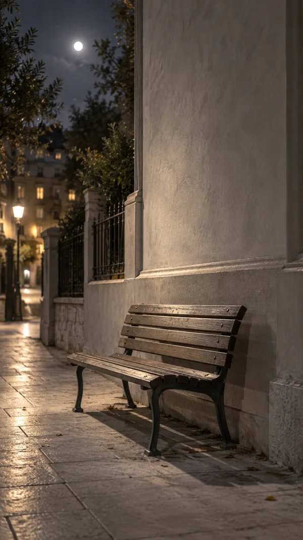 An empty weathered wooden-and-metal bench sits against a tall pale stone wall on a nighttime city sidewalk, with trees, a wrought-iron fence, lit windows…