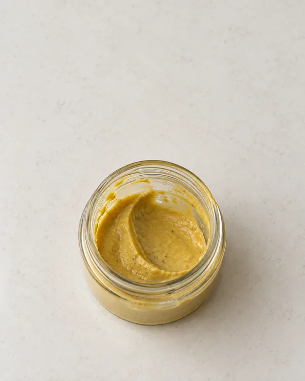 Direct overhead view of an open, clear glass jar partly filled with golden mustard sauce, set below centre on a lightly mottled off-white surface.