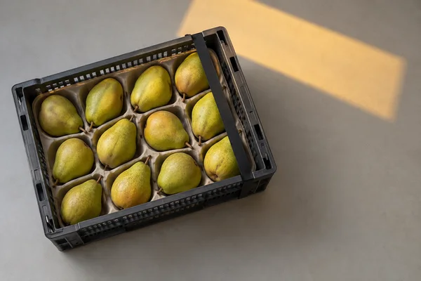 A charcoal-grey ventilated crate filled with yellow-green pears in individual pulp pockets sits on a pale-grey tabletop, viewed from above.