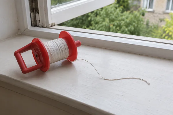 A red plastic cord reel rests diagonally on a pale windowsill beside an open casement, with white cord trailing in a gentle curve across the sill and greenery…