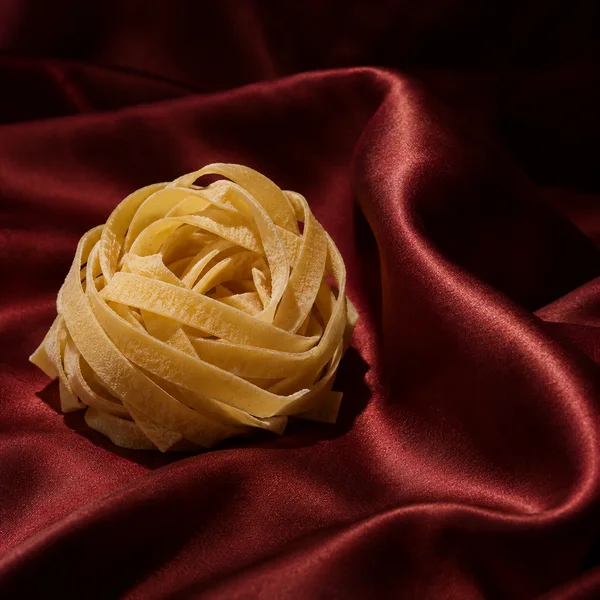 A pale yellow tagliatelle nest rests on deep red satin, with a raised glossy fabric fold to the right and dark space in the upper-right corner.