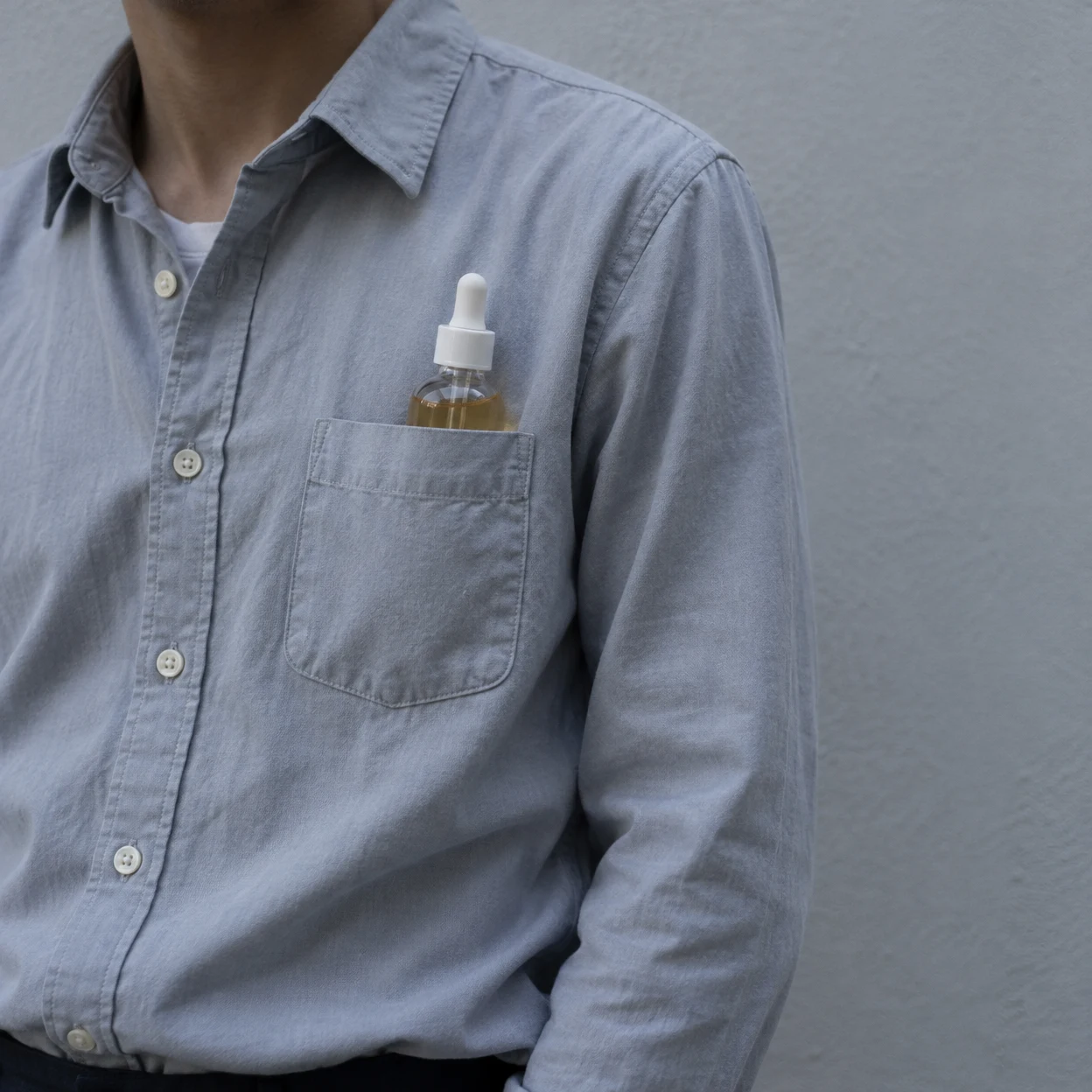 Cropped portrait of an adult wearing a pale blue-grey button-up shirt, with a clear dropper bottle of amber liquid tucked into the chest pocket against a plain…