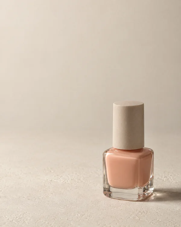 A clear rounded-square bottle filled with muted peach nail polish sits in the lower-right of a warm ivory textured surface.