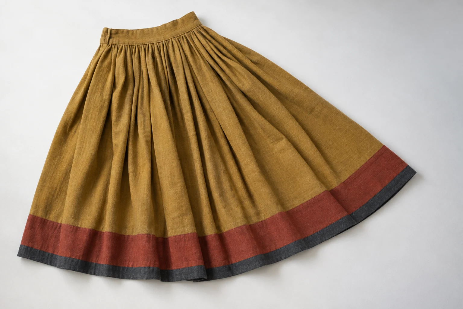 Overhead studio view of an ochre pleated wool skirt on an off-white surface, with a broad rust-red hem band and narrow charcoal edging.