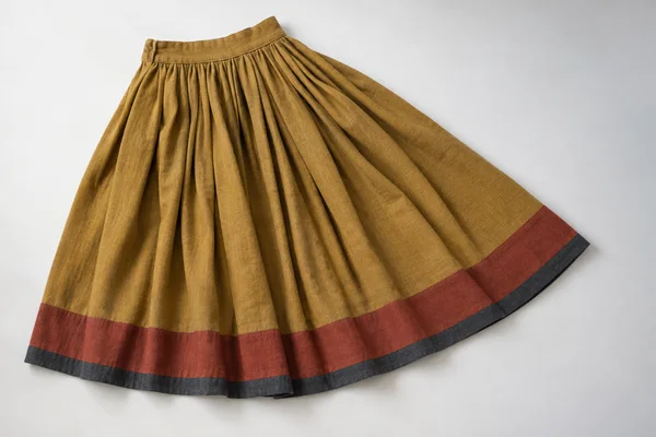 Overhead studio view of an ochre pleated wool skirt on an off-white surface, with a broad rust-red hem band and narrow charcoal edging.