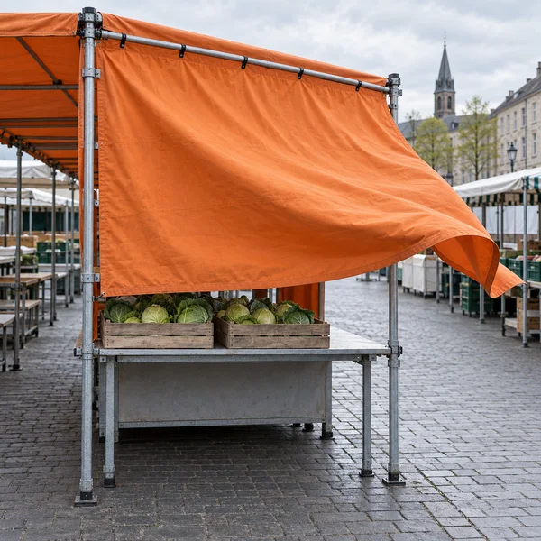 A galvanized market stall stands on a cobbled street, with two wooden crates of green cabbages beneath a large orange canvas curtain billowing outward in the…
