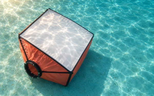 A coral-sided square softbox lies diagonally on a turquoise tank floor, its white diffuser facing upward with water patterns across it; a black circular…