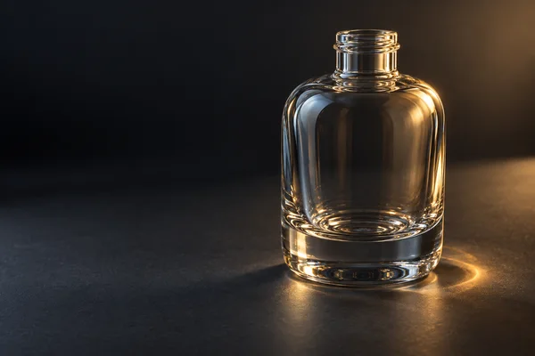 A clear, thick-based glass bottle stands on a dark charcoal surface, lit with warm gold highlights and a bright crescent of light beside its base; the bottle…