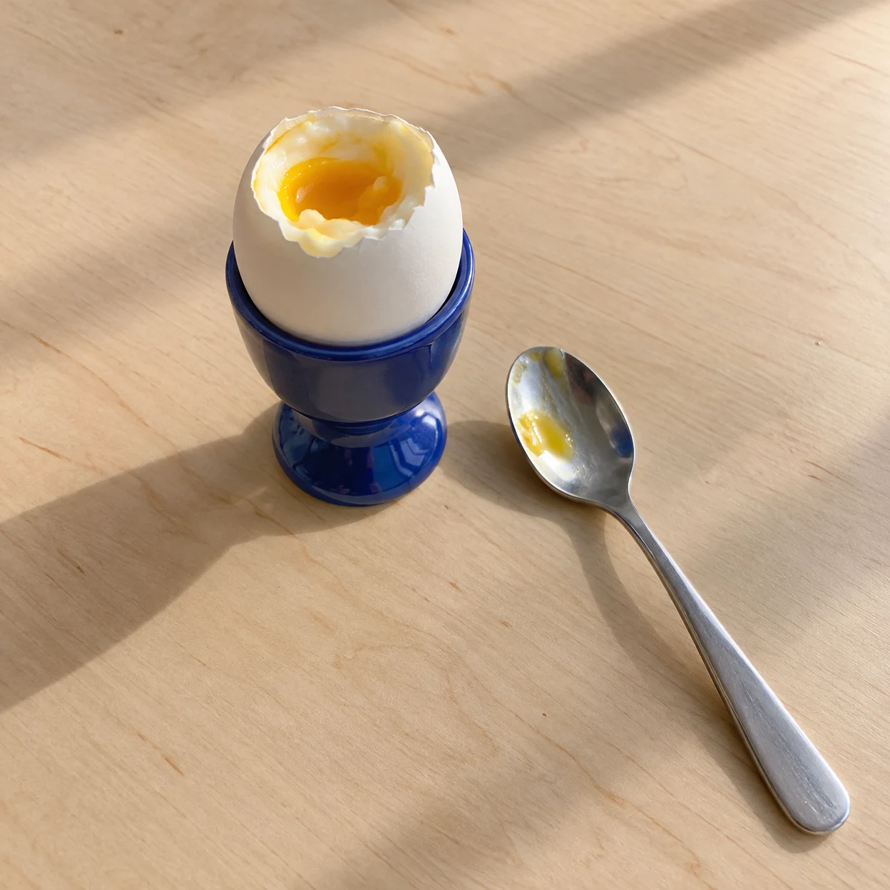 A white soft-boiled egg with its top unevenly opened sits in a cobalt-blue ceramic eggcup on a pale wood table.