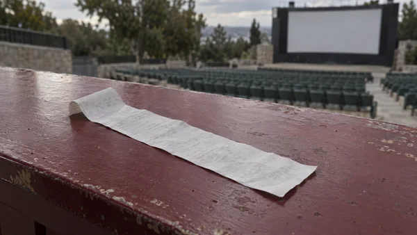 A long, faintly printed receipt lies diagonally across a weathered burgundy counter, with rows of empty seats and a blank outdoor cinema screen softly blurred…
