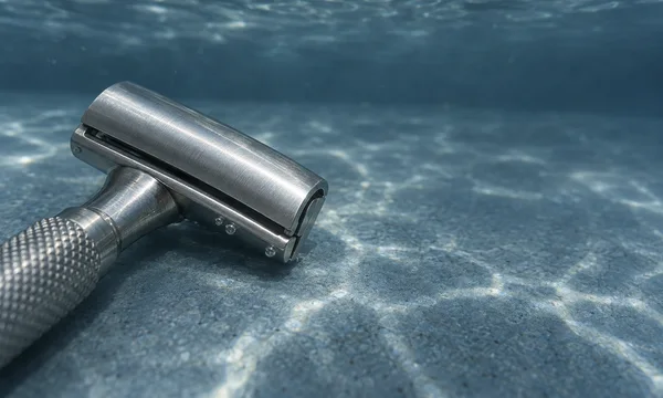 A stainless-steel safety razor lies diagonally on a blue-grey submerged floor, its large brushed-metal head in sharp focus and knurled handle extending out of…