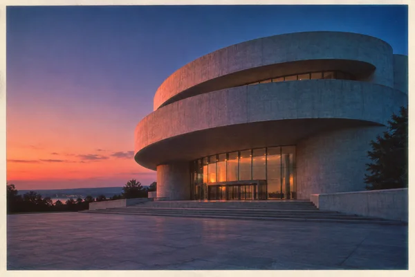 A sweeping, tiered concrete rotunda with a recessed glass entrance stands across an empty stone forecourt at dusk.