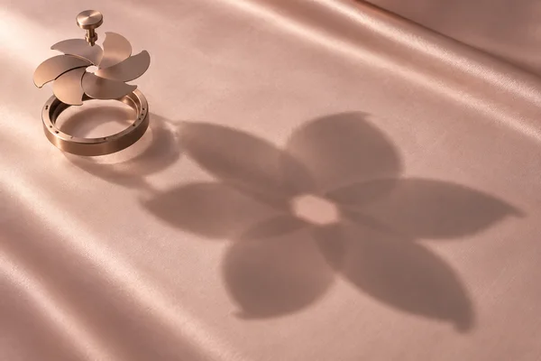A rose-gold mechanical flower shutter is shown in separated layers above pale blush satin, casting a large, softly edged flower-like shadow toward the lower…