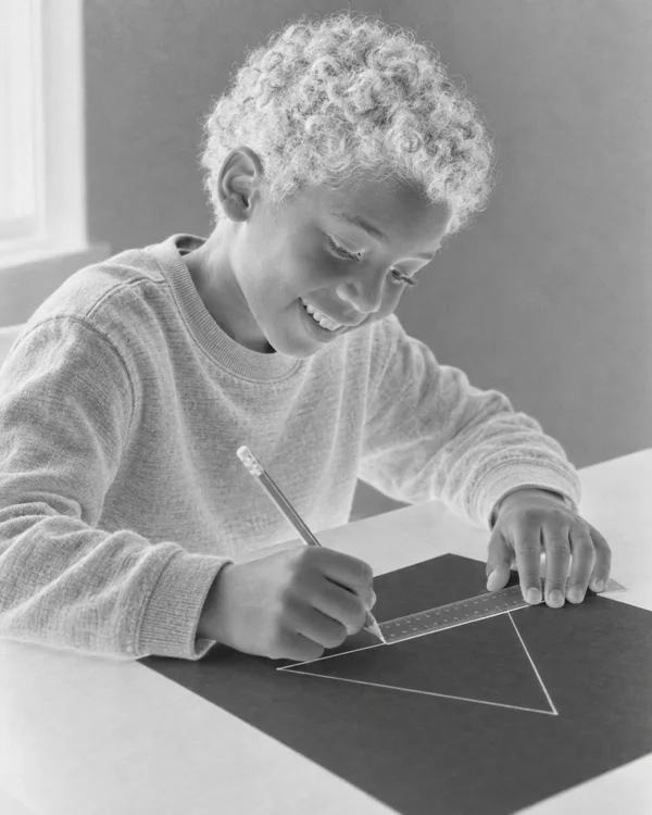 A smiling child leans over a dark worksheet in a high-key grayscale negative-style image, using a pencil and ruler to draw a large outlined triangle.