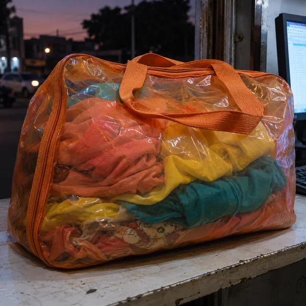 A translucent orange zippered carryall packed with coral, yellow and turquoise clothing rests on a worn counter at a street-facing kiosk at dusk; a computer…