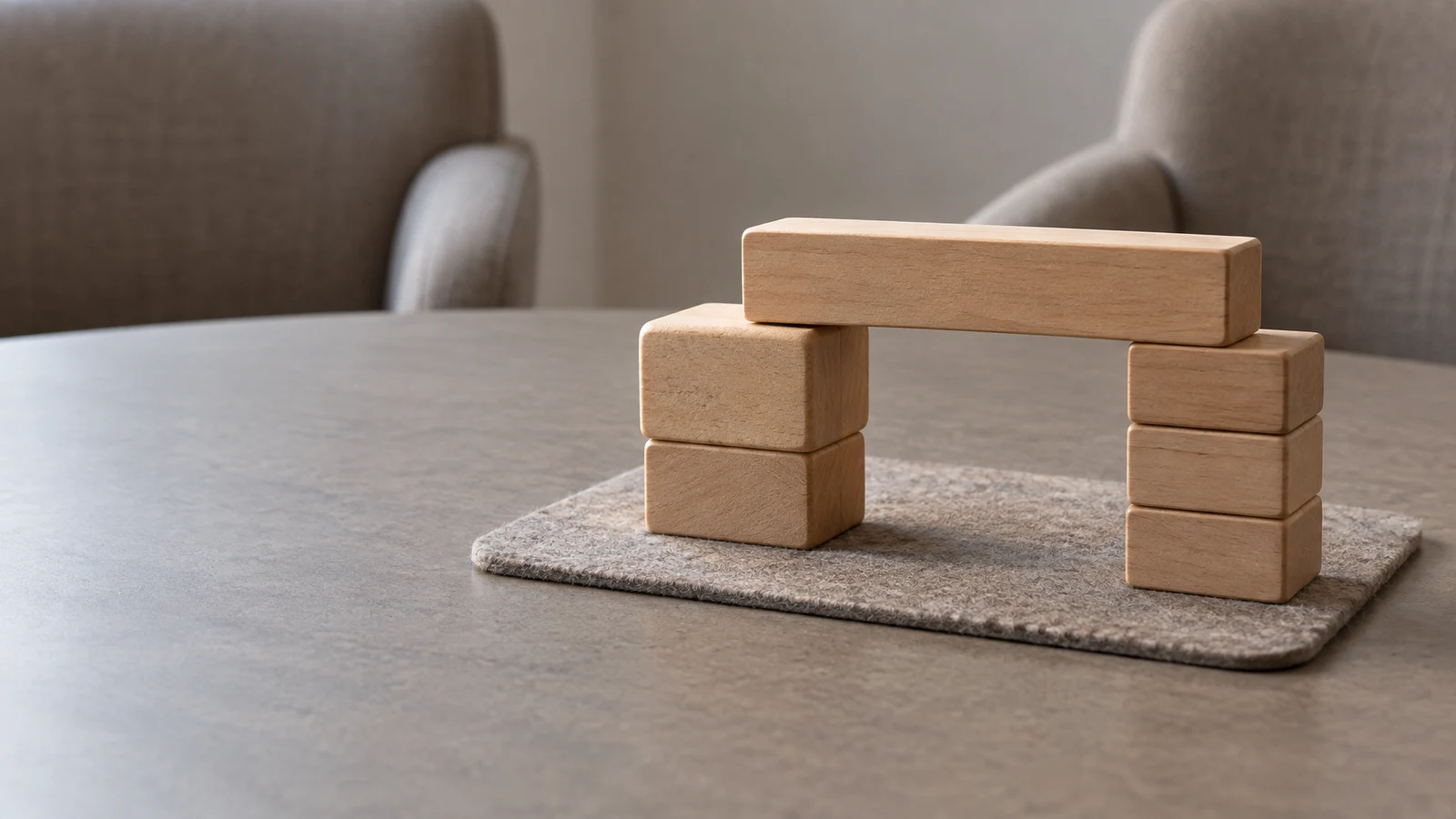 A long wooden block bridges two stacks of wooden blocks on a small oatmeal-grey felt mat, with two blurred upholstered chairs in the background and open grey…