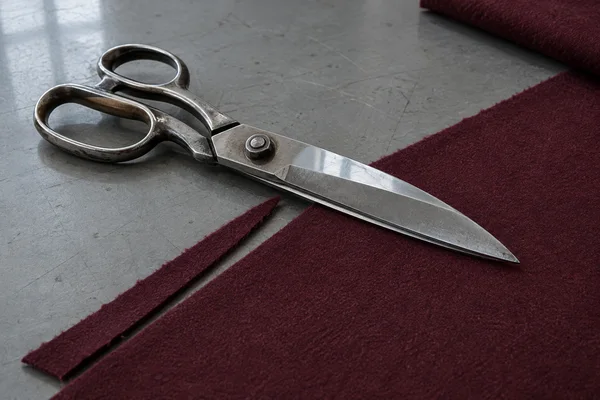 Large steel tailor’s shears lie diagonally across burgundy wool on a scratched grey worktable, beside a narrow strip of cut fabric.