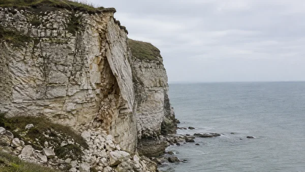 A pale layered sea cliff fills the left side of the frame, with a large thin sheet of rock peeling outward above a pile of broken fragments.