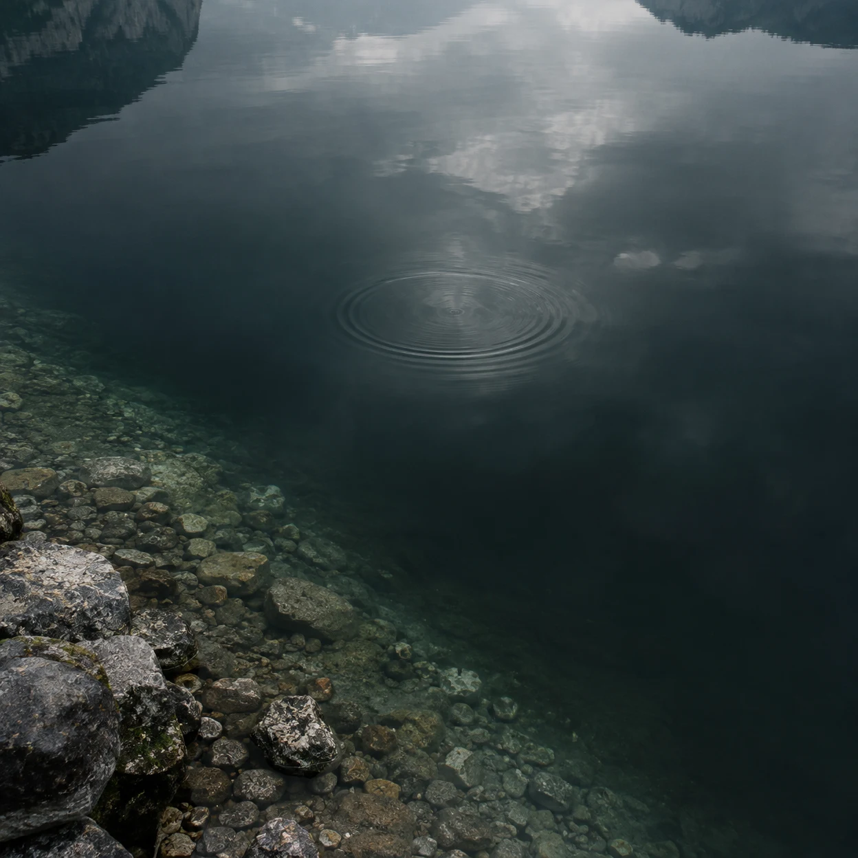 A high-angle view over a dark slate-green lake, with a broad bed of submerged pebbles and shoreline rocks in the lower left, cloud reflections on the water…