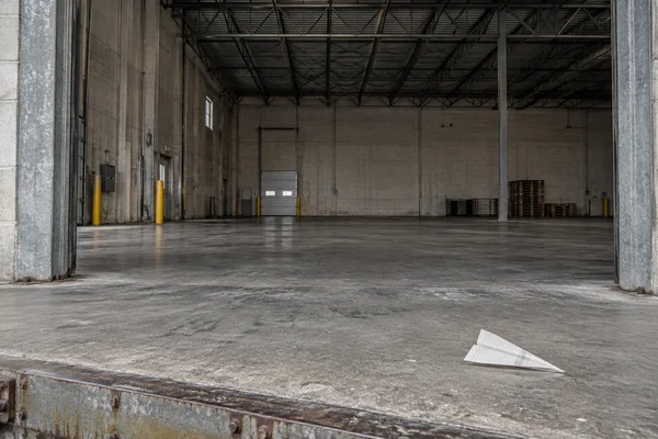 A small folded paper airplane made from a ruled, blank form rests on the worn concrete loading dock in front of a large open warehouse bay, with steel roof…
