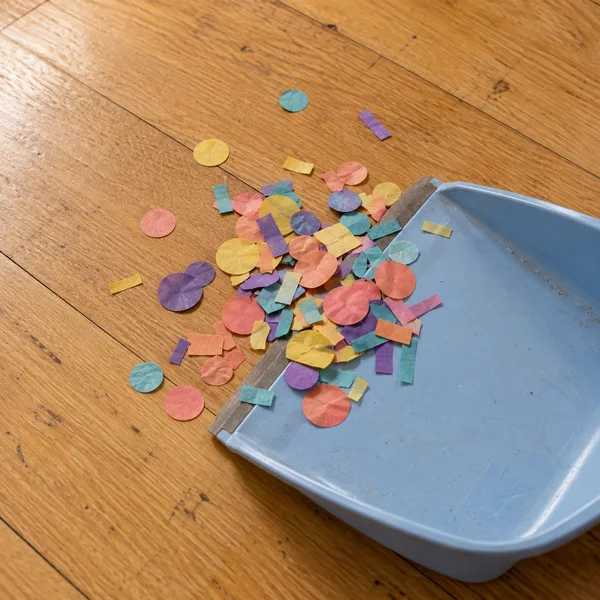 An irregular heap of coral, yellow, violet and turquoise paper circles and strips gathers along the edge of a pale-blue plastic dustpan on a honey-coloured…