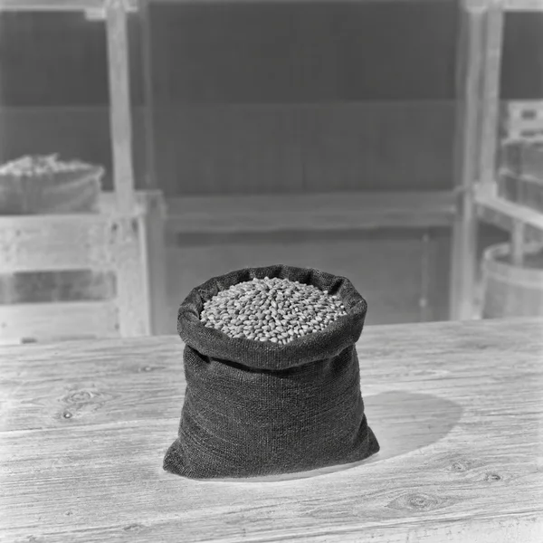 A dark fabric sack filled with small pale grains sits on a light wooden counter in a softly blurred market-stall setting; the monochrome image has a…