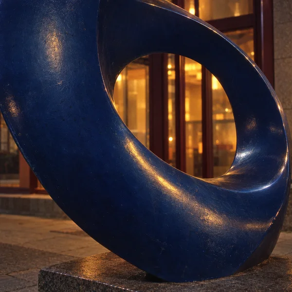 A large deep-blue steel ribbon sculpture curls around a complete oval opening, standing on a granite plinth beside paved ground.