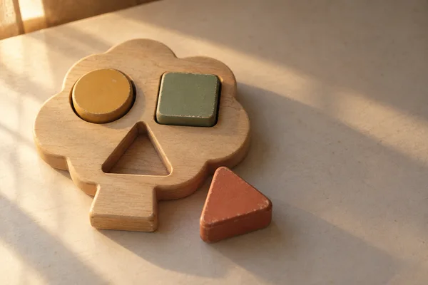 A worn wooden tree-shaped sorter sits left of center on a pale tabletop, with a mustard circle and sage square in their recesses and a terracotta triangle…