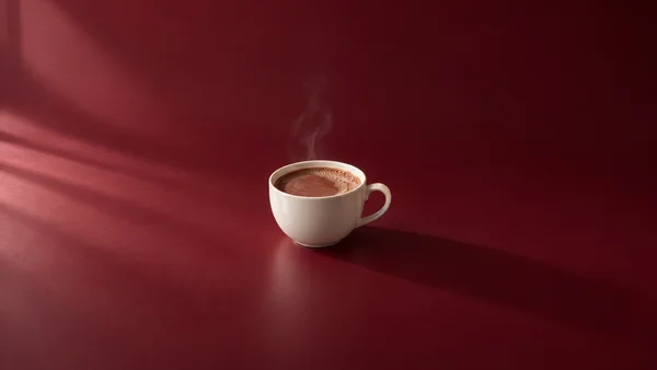 An ivory porcelain cup filled with brown hot chocolate sits on a deep red studio floor, with a thin wisp of steam rising above it and a long shadow extending…
