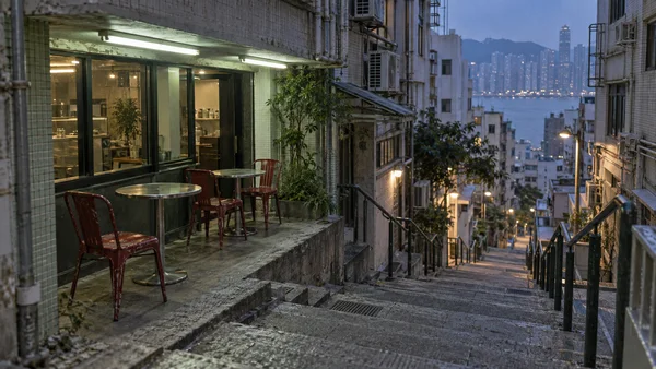 An empty cafe terrace with stainless-steel tables and dark-red metal chairs sits beside a steep Hong Kong stairway at dusk, with tiled buildings, utility…