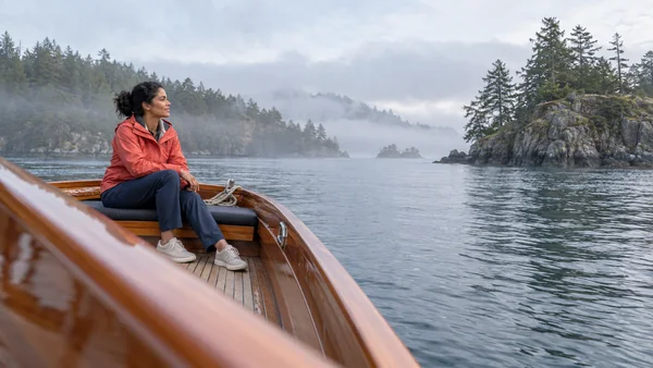 A woman in a coral jacket and navy trousers sits on a cushioned wooden launch bench, looking across silver-gray water toward wooded islands softened by sea fog.
