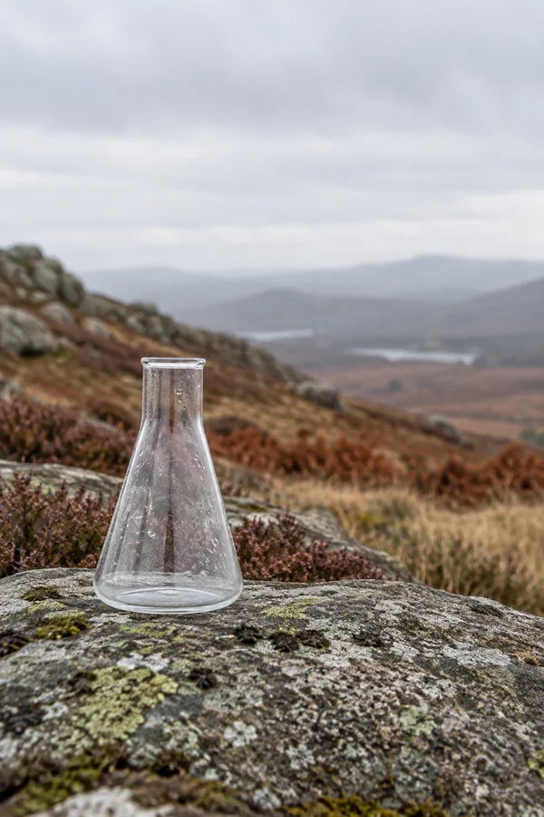 A clear Erlenmeyer flask dotted with moisture stands on a lichen-covered granite boulder, with muted heather and bracken, a hazy upland landscape, and a broad…