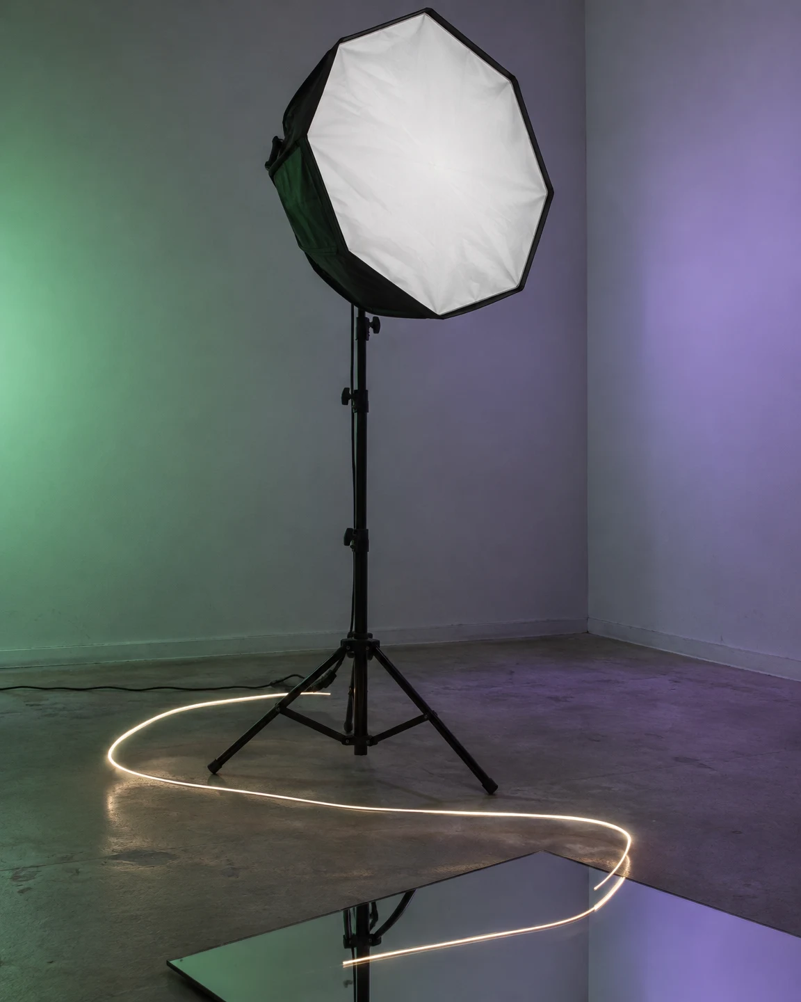 An octagonal white softbox on a black stand sits in a bare studio corner washed green on the left and violet on the right.