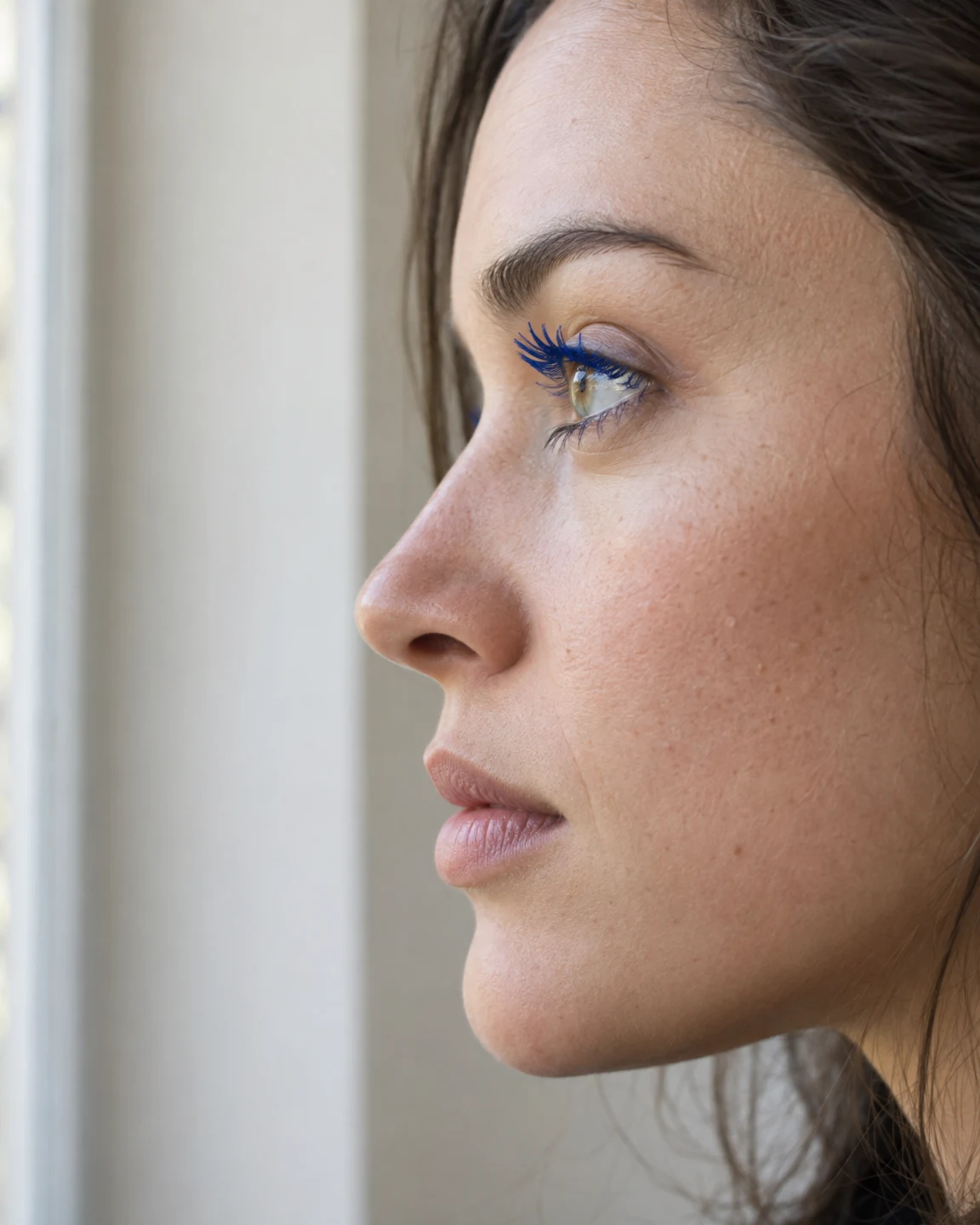 A close side-profile portrait of an adult with dark hair looking toward a bright window at the left.