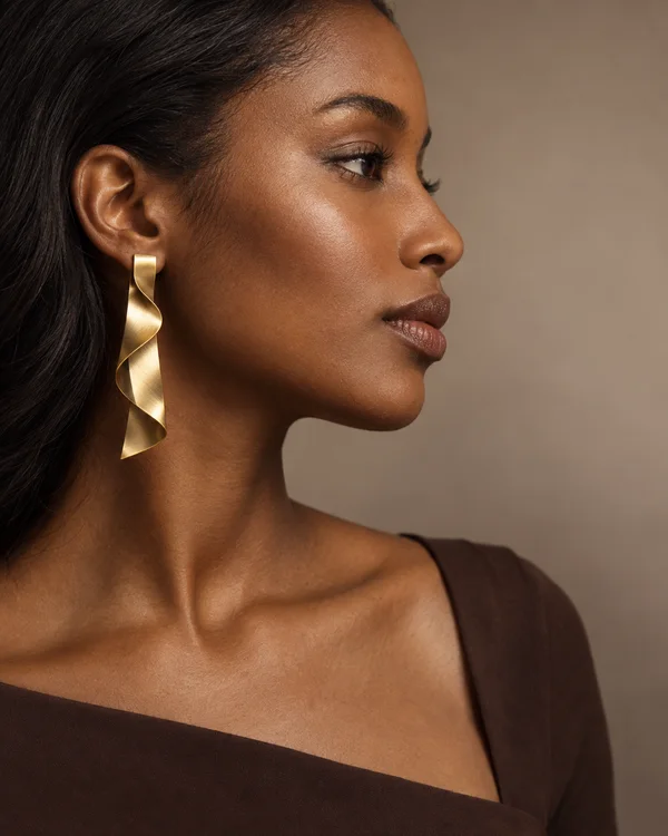 A woman with dark hair swept behind her ear wears a long folded gold ribbon earring, shown in side profile against a warm taupe studio background.