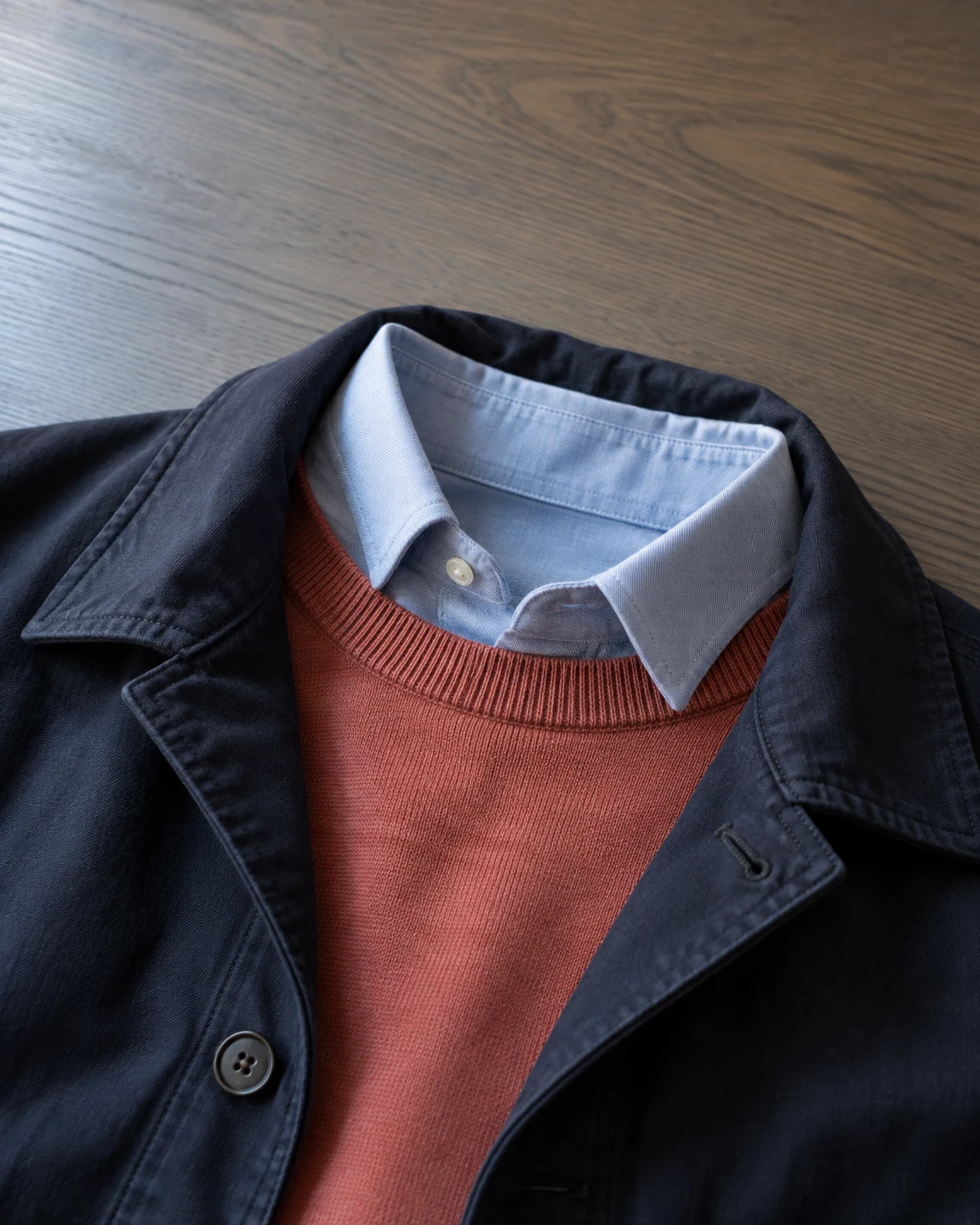 An open dark navy jacket lies over a coral ribbed sweater and pale blue collared shirt on an ash-wood tabletop, viewed closely from above.