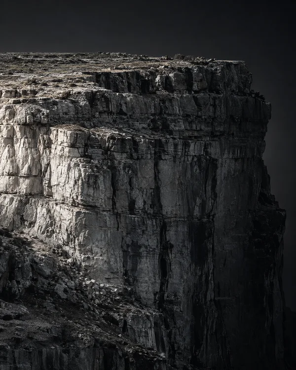 A pale, horizontally layered cliff face with deep vertical fissures fills most of the frame, lit from the left against a charcoal-black background.