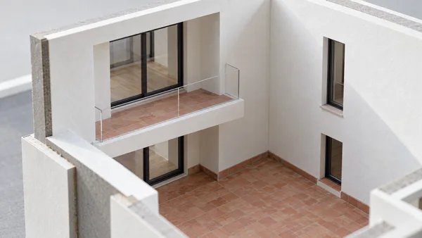 An elevated close view of a white architectural model showing a terracotta-tiled courtyard, a recessed balcony with a clear balustrade, and dark-framed doors…