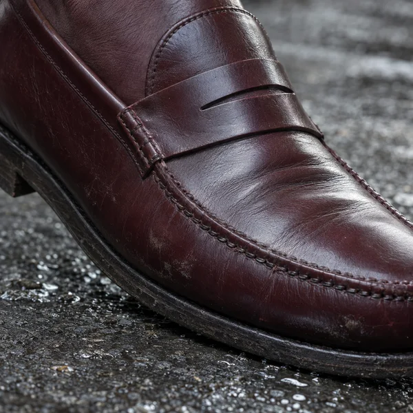 Extreme closeup of a dark burgundy leather penny loafer, showing its slit strap, curved stitched seam and creased vamp above damp, textured pavement; both ends…