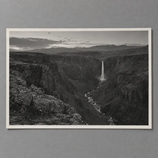 A black-and-white landscape photograph of a rocky gorge and narrow waterfall lies as a bordered paper print on a smooth gray surface, with distant hills and…