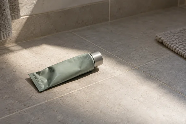 A muted sea-green cream tube with an attached silver cap lies diagonally on sunlit bathroom tiles, casting a defined shadow; a towel and textured bath mat…