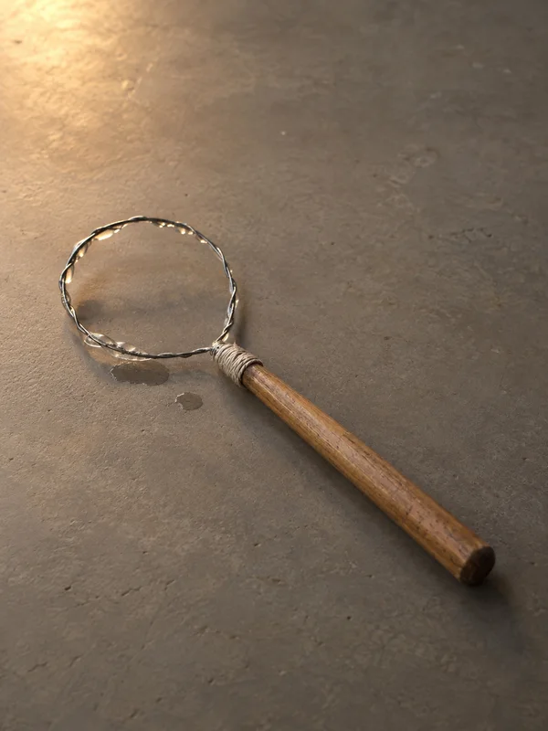 A handmade bubble wand lies diagonally on a warm grey tabletop, with a reflective wire loop, a cotton-wrapped wooden handle, and two small wet marks nearby.