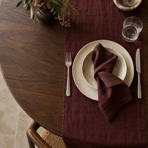 Overhead view of a cream stoneware plate and shallow bowl with a burgundy linen napkin, silver cutlery and a water glass on a dark walnut table with a matching…