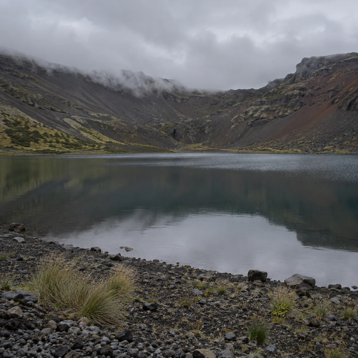 A still slate-gray lake lies within a steep volcanic crater, reflecting the cloudy sky faintly.