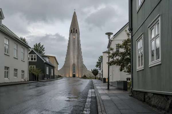 A tall stepped concrete church rises at the end of a rain-darkened street, framed by low buildings on the left and a large corrugated-metal building on the…